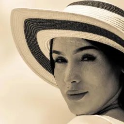 Portret kobiety w kapeluszu z szerokim rondem, w sepii, z delikatnym uśmiechem i spojrzeniem przez ramię.