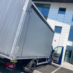 Niebieski bus MAN z otwartymi drzwiami na tle nowoczesnego budynku biurowego z szarym plandekowym nadwoziem.