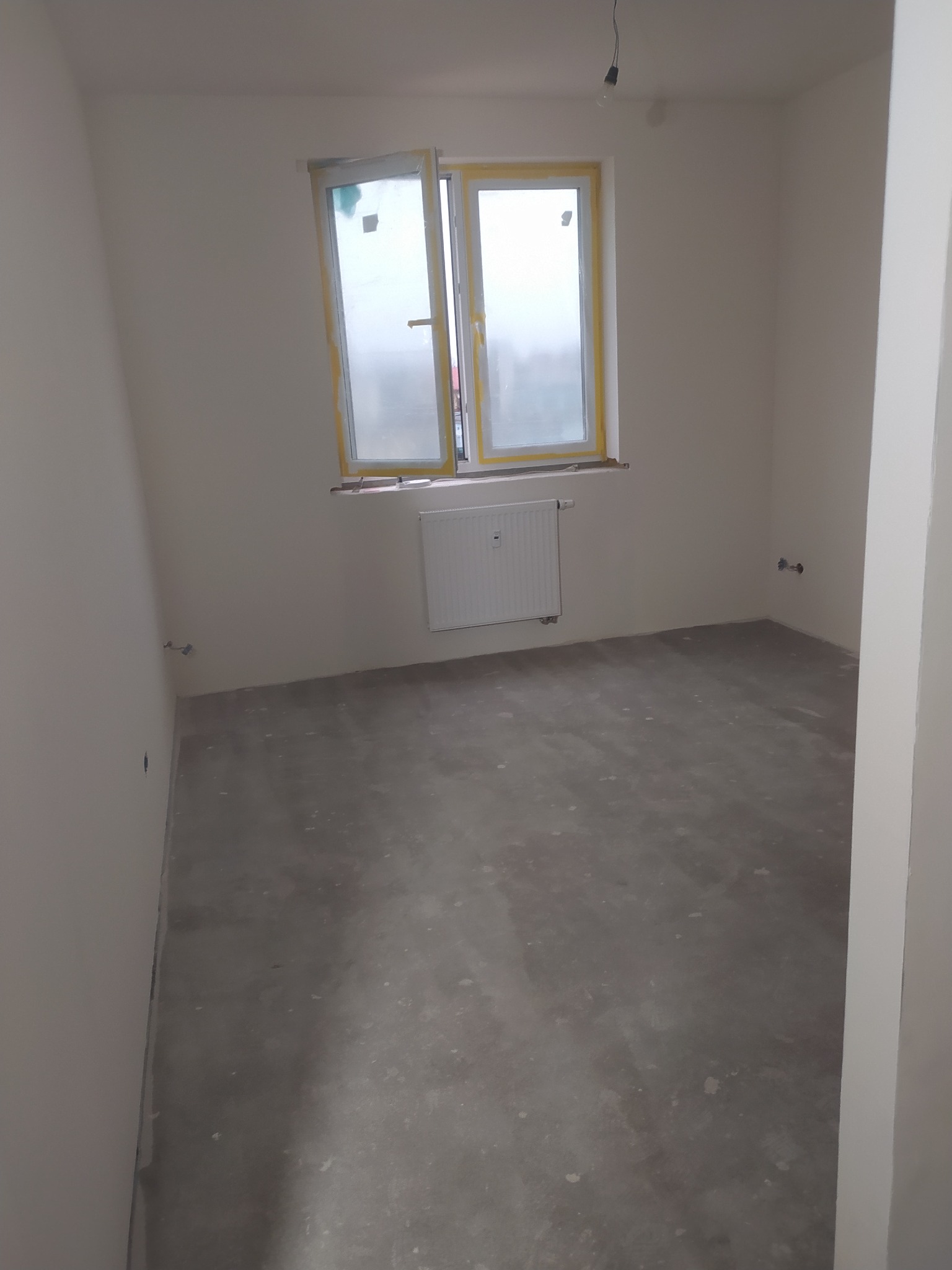 Pusty pokój w trakcie remontu: ściany po szpachlowaniu i malowaniu na biało, surowa betonowa podłoga, okno z żółtą folią ochronną na ramie, grzejnik pod oknem, widoczne wyprowadzenia instalacji...
