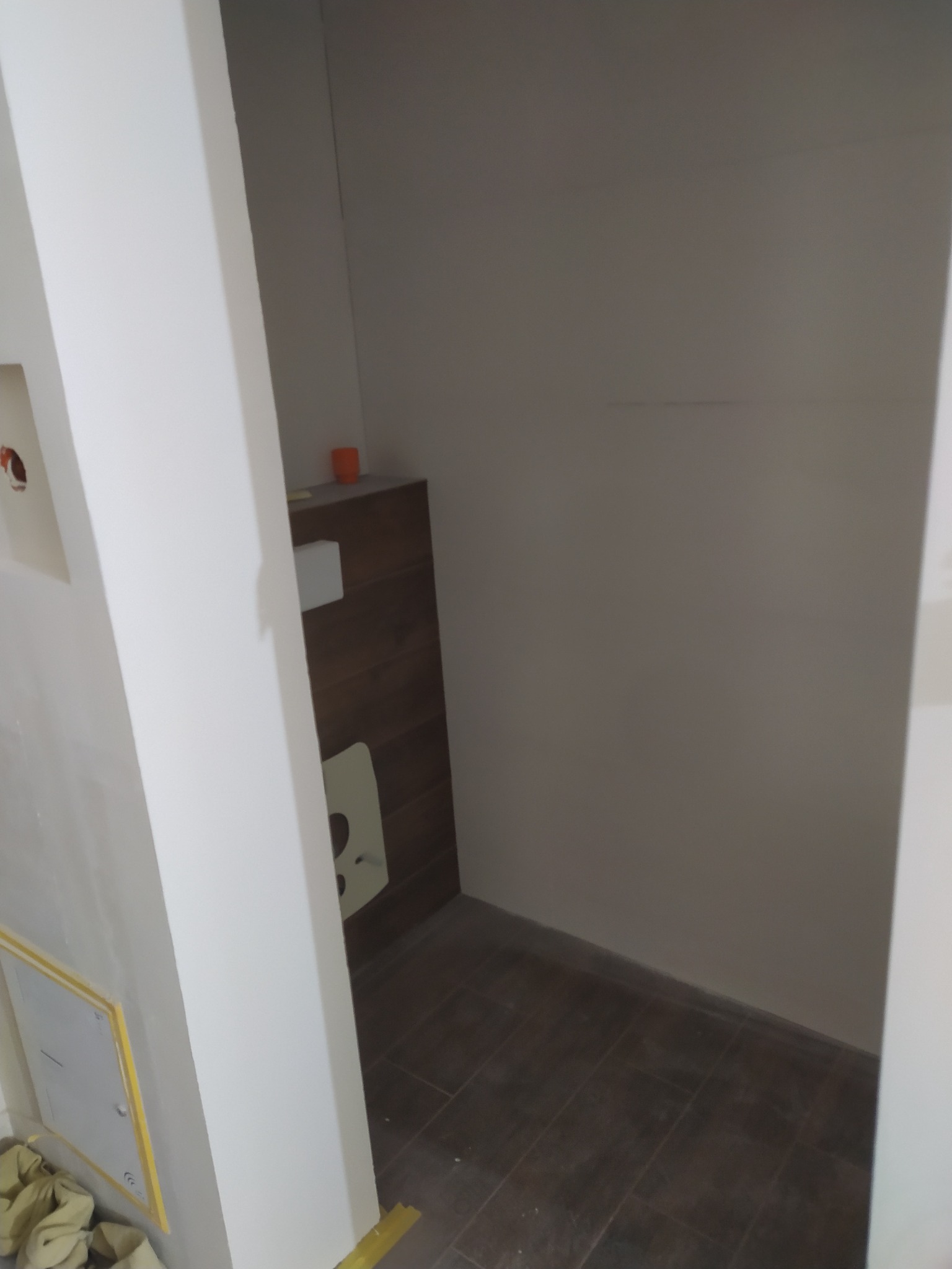 Wnęka w trakcie wykańczania: brązowe płytki podłogowe, fragment obudowy stelaża podtynkowego z drewnopodobnymi panelami, widoczne przygotowania do malowania ścian, zabezpieczona żółtą taśmą maskującą.