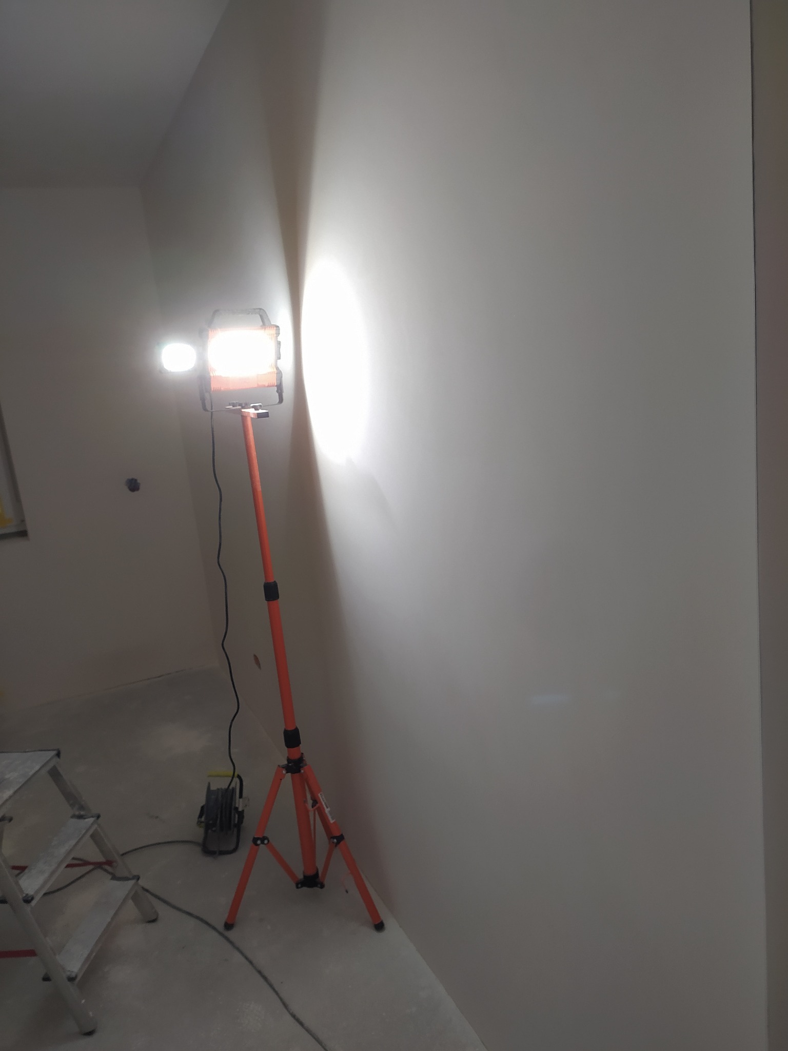 Świeżo pomalowana ściana oświetlona lampą roboczą na statywie; widoczna drabina malarska i zwinięty przedłużacz.