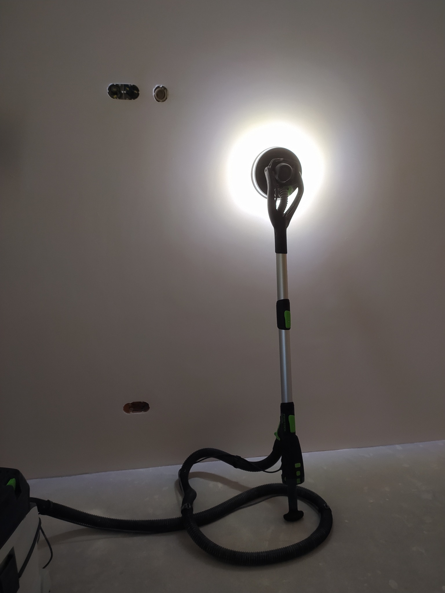 Szlifierka przegubowa z odkurzaczem do gładzi, oświetlająca obrabianą powierzchnię sufitu z przygotowanymi otworami na instalacje.