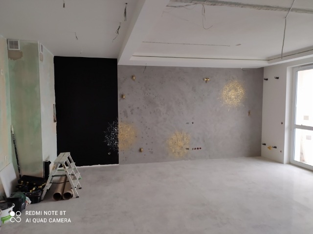 Wnętrze w trakcie prac wykończeniowych: szpachlowana ściana z dekoracyjnymi złotymi wzorami, czarna ściana, drabina i narzędzia malarskie w rogu pomieszczenia.