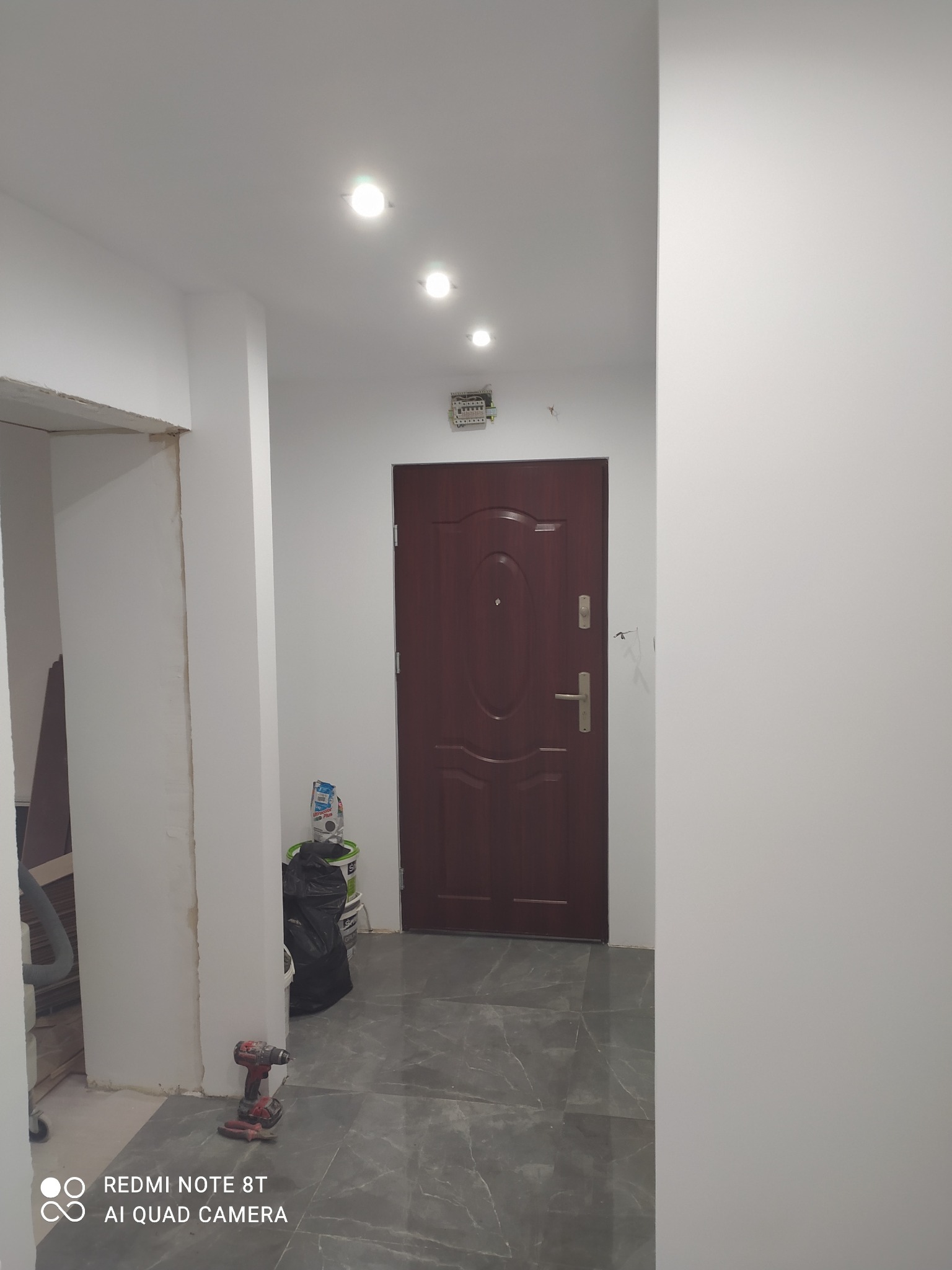 Korytarz po remoncie: szare płytki podłogowe, pomalowane na biało ściany, brązowe drzwi wejściowe. Widoczny wiertarko-wkrętak akumulatorowy i wiadro z farbą.