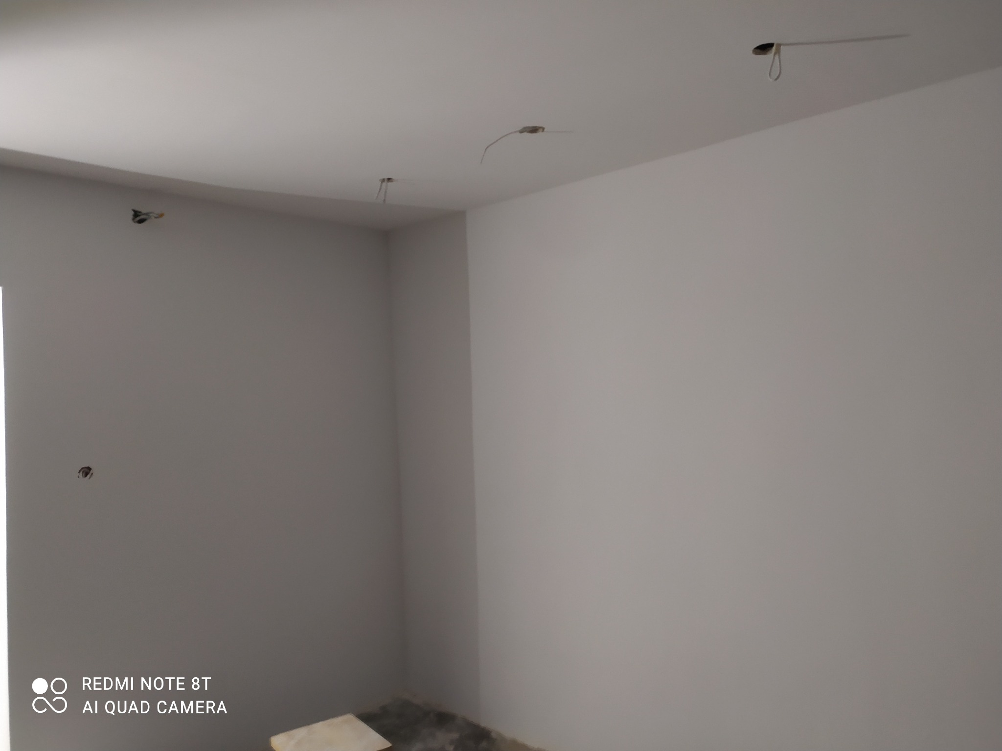 Pomalowane na szaro wnętrze pokoju w trakcie prac wykończeniowych, widoczne wystające kable ze ścian i sufitu, fragment podłogi z resztkami materiałów budowlanych.