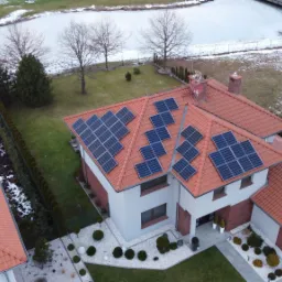 Widok z lotu ptaka na dom jednorodzinny z panelami słonecznymi na dachu z czerwonej dachówki, obok zamarznięte jezioro i dwa białe samochody zaparkowane w garażu.