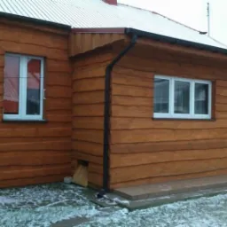 Dom z drewnianą elewacją i białymi oknami, widoczne rynny i fragment zadaszenia z drewnianą konstrukcją kratową, na ziemi resztki śniegu.