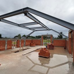 GASE INVEST GROUP SP Z O.O. - Wnętrze budowy domu z czerwonej cegły z metalową konstrukcją dachu i mokrą podłogą. Widoczne palety z cegłami i niezadaszony otwór. Pochmurne niebo w tle.