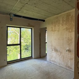 GASE INVEST GROUP SP Z O.O. - Wnętrze surowego pokoju z oknem, widoczne ślady po tynku na ścianach, betonowy sufit z instalacją. Widok na zieleń za oknem. Fragment ściany z cegły po prawej.