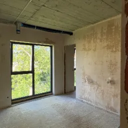 Wnętrze surowego pokoju z oknem, widoczne ślady po tynku na ścianach, betonowy sufit z instalacją. Widok na zieleń za oknem. Fragment ściany z cegły po prawej.