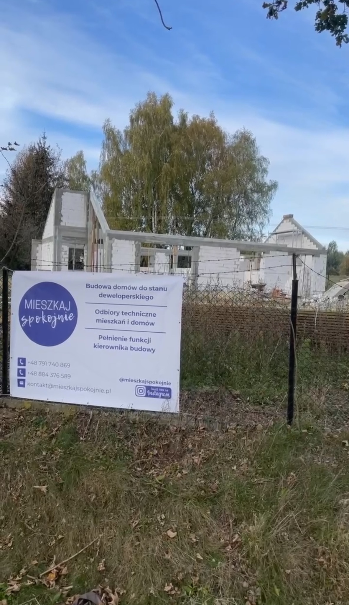 Niedokończony dom za ogrodzeniem z reklamą usług: budowa domów, odbiory techniczne, kierownik budowy, z danymi kontaktowymi i logo firmy.