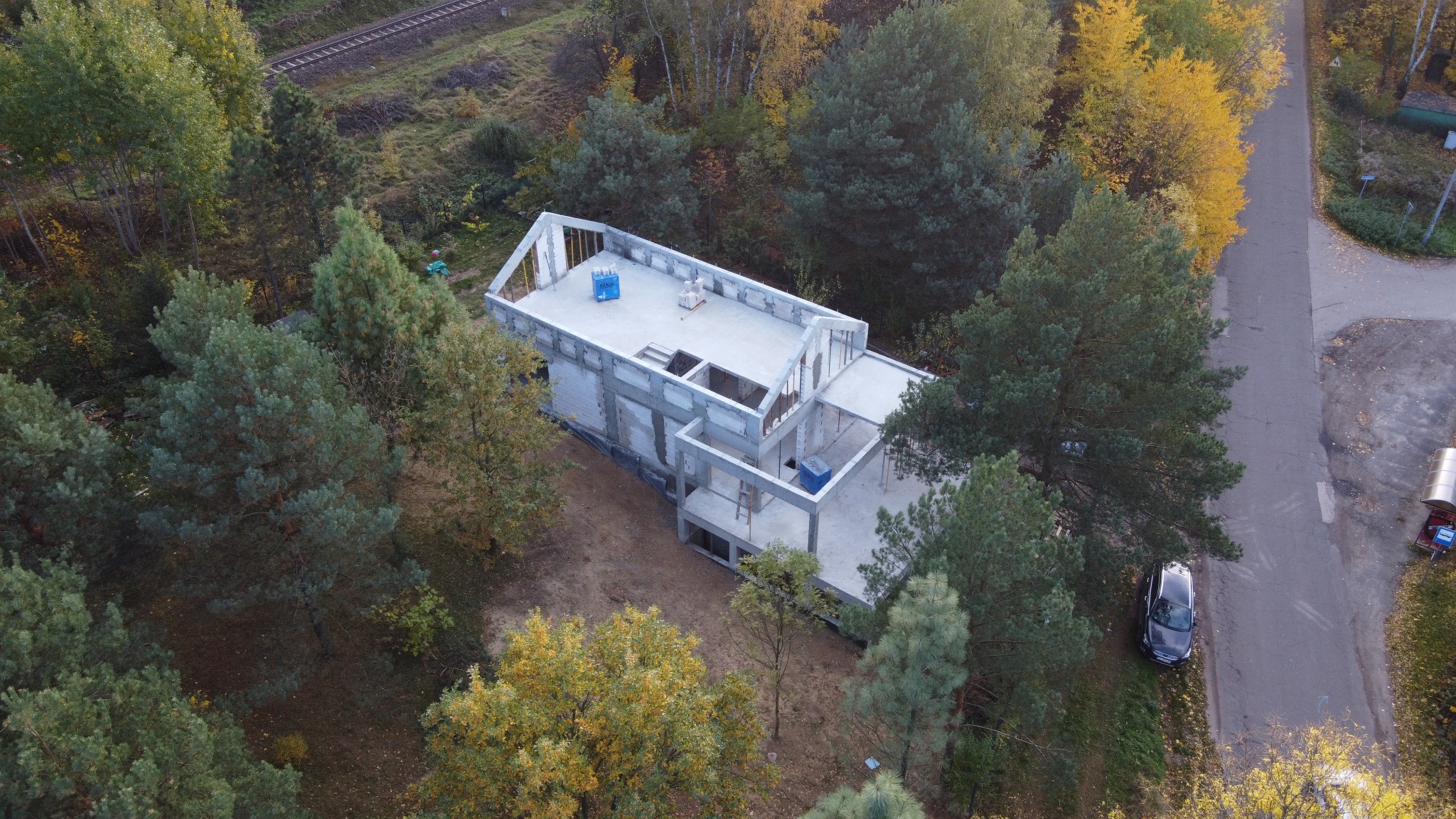 Zdjęcie z drona: surowy, dwukondygnacyjny dom w trakcie budowy, otoczony jesiennym lasem, z widoczną konstrukcją ścian i stropów. W tle tor kolejowy i droga.