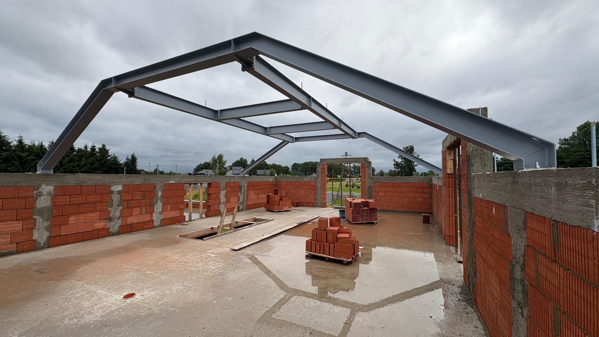 Wnętrze budowy domu z czerwonej cegły z metalową konstrukcją dachu i mokrą podłogą. Widoczne palety z cegłami i niezadaszony otwór. Pochmurne niebo w tle.