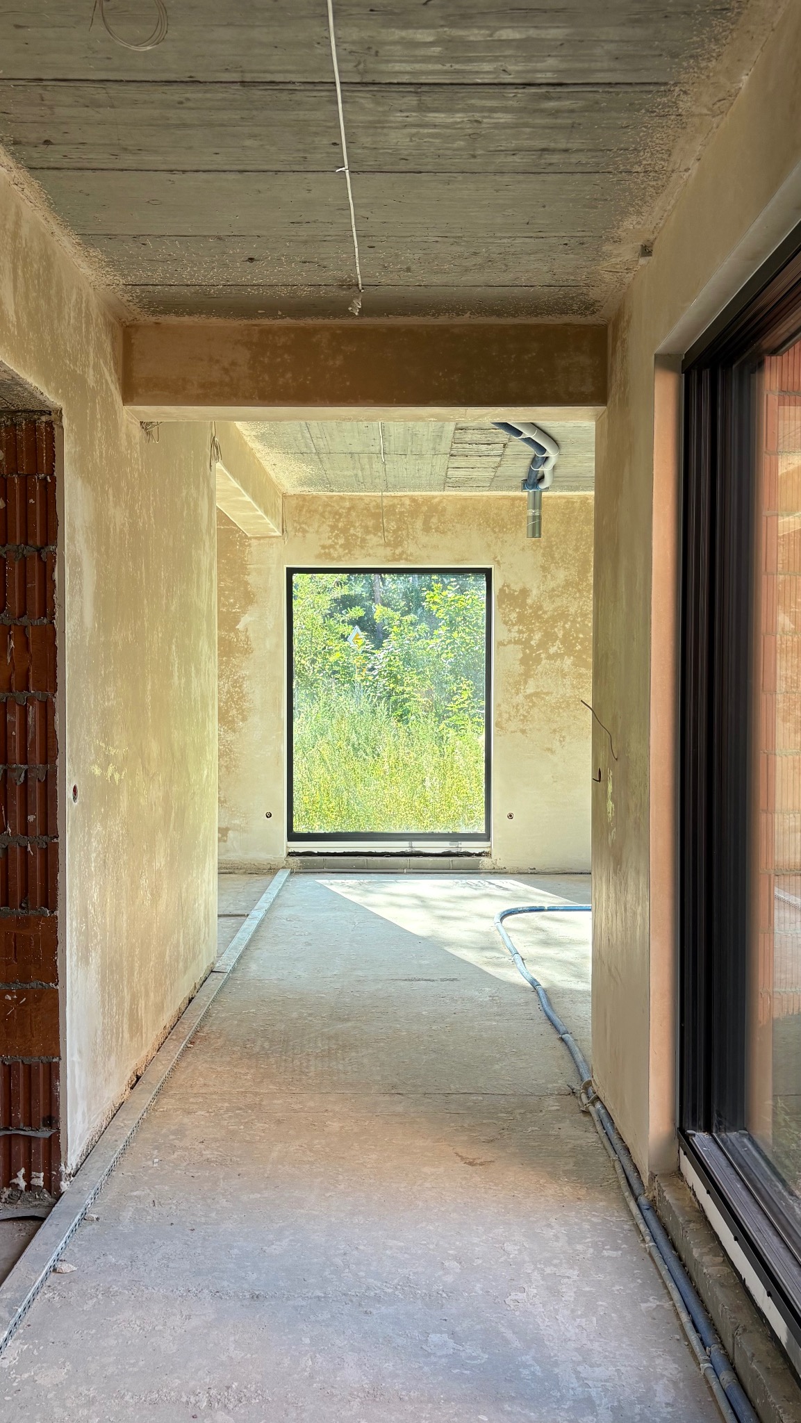 Wnętrze surowego domu w trakcie budowy: betonowa podłoga, okno z widokiem na zieleń, ściany w tynku, instalacje. Widok z korytarza.
