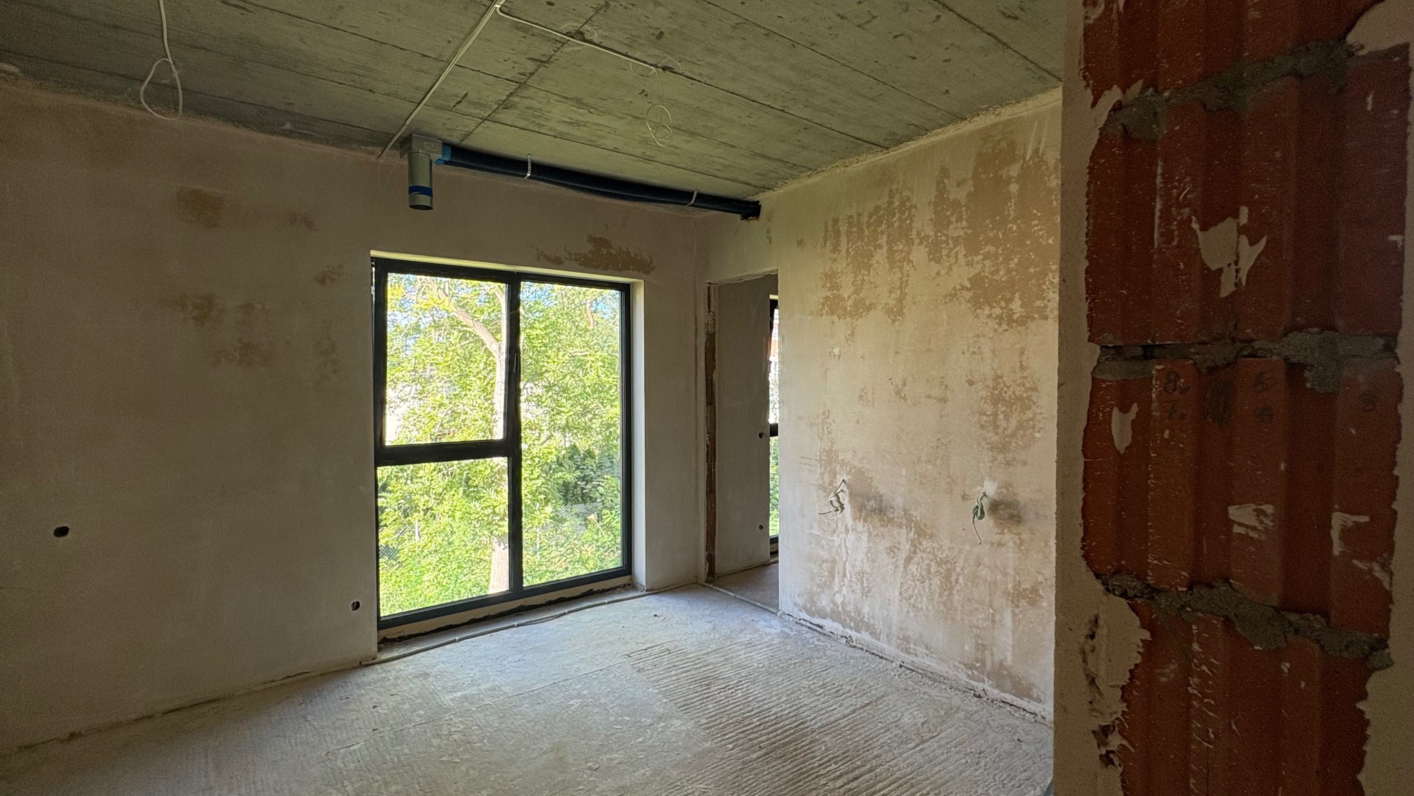 Wnętrze surowego pokoju z oknem, widoczne ślady po tynku na ścianach, betonowy sufit z instalacją. Widok na zieleń za oknem. Fragment ściany z cegły po prawej.
