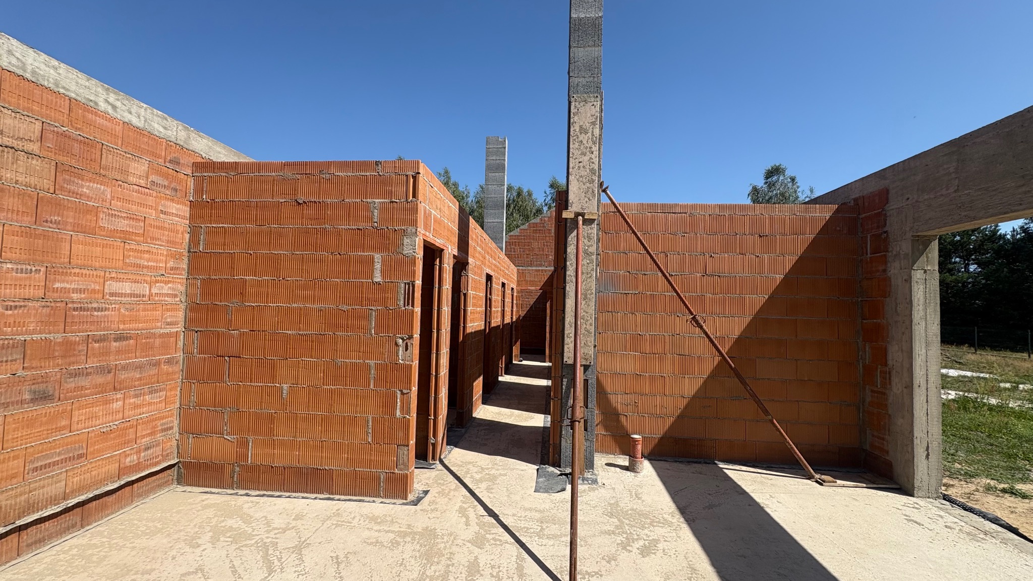 Wnętrze domu w trakcie budowy z czerwonej cegły, widoczne ściany z otworami na drzwi, betonowe słupy i podpory. Jasne, słoneczne oświetlenie.