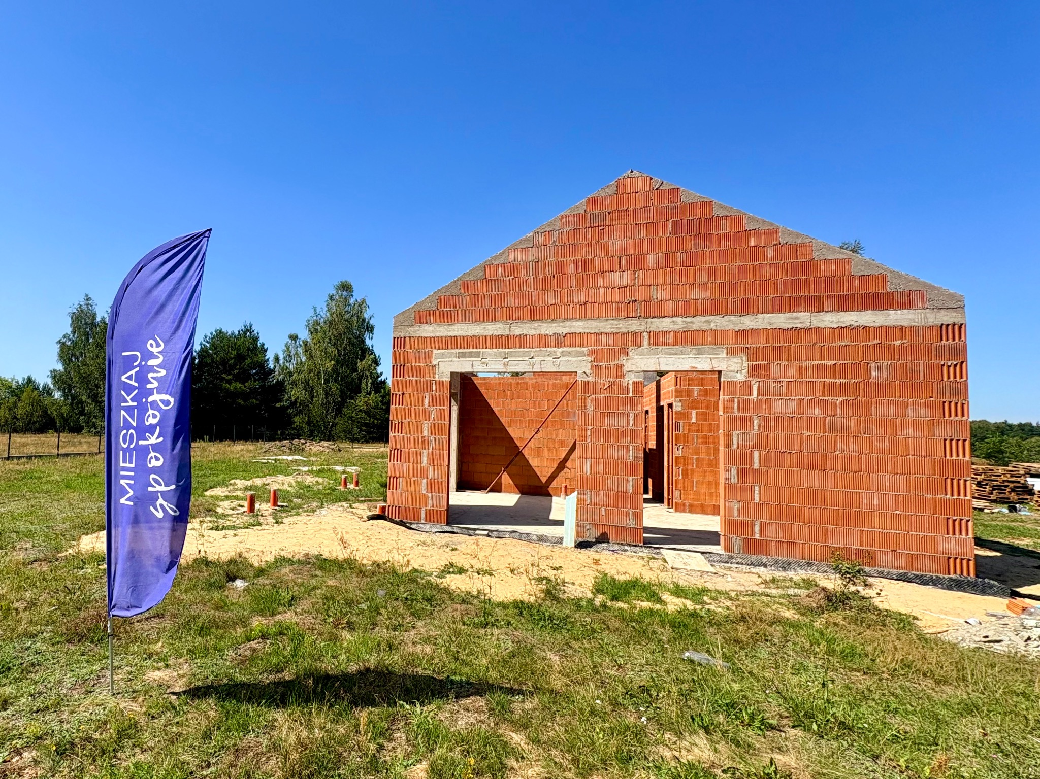 Wznoszenie ścian parterowego domu z czerwonej cegły ceramicznej. Na pierwszym planie flaga z napisem 'Mieszkaj Spokojnie'.