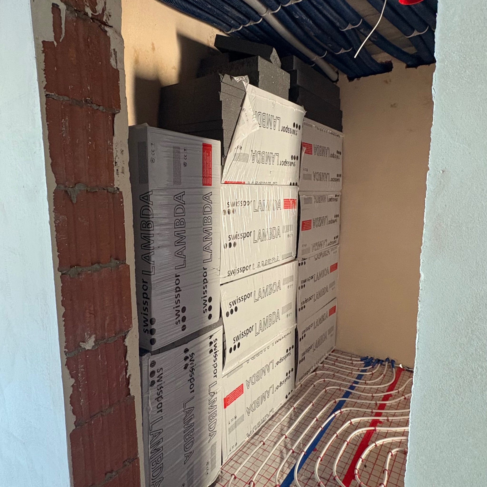 Pakiety izolacji Swisspor Lambda obok instalacji ogrzewania podłogowego w surowym pomieszczeniu z widoczną cegłą. Etap prac wykończeniowych.