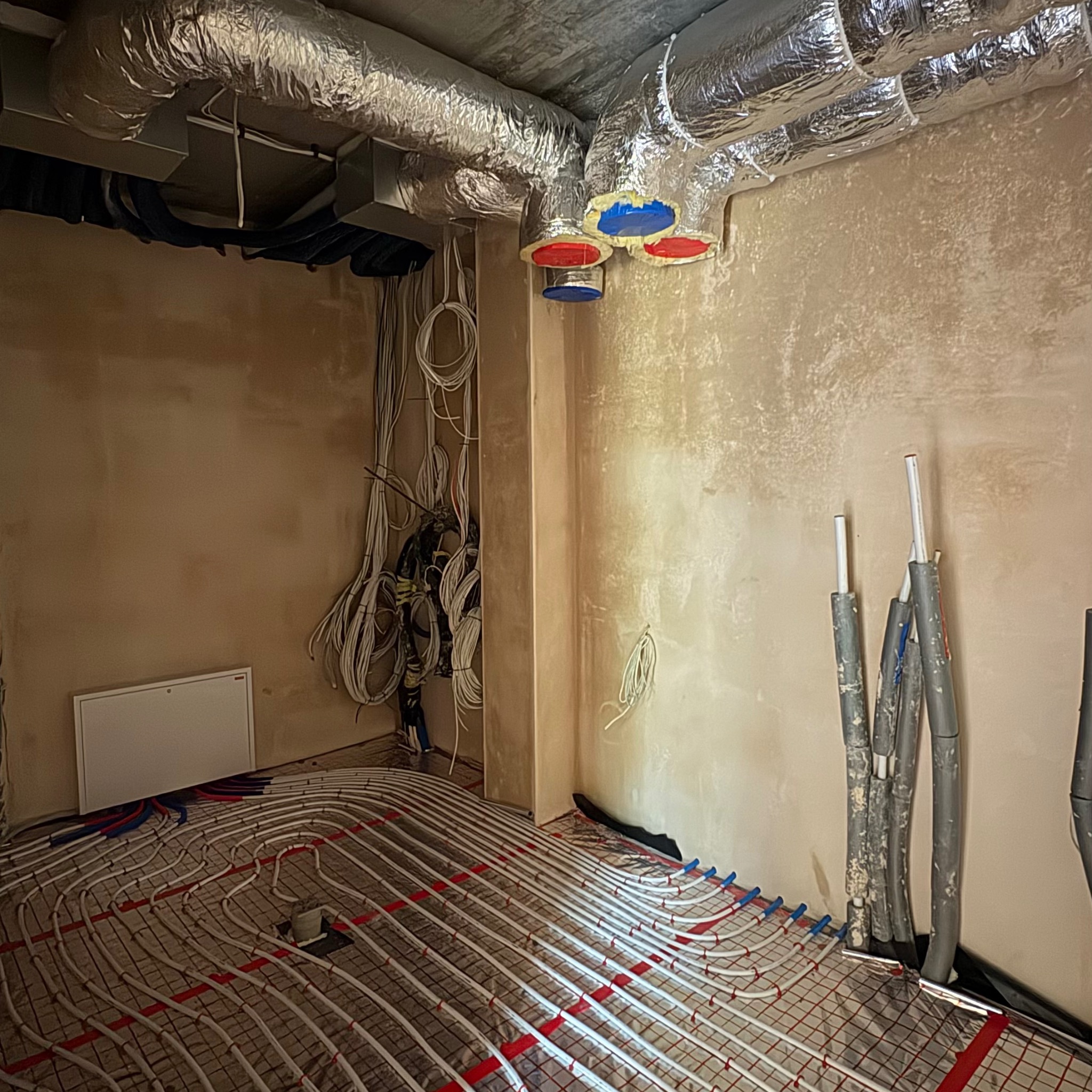 Instalacja ogrzewania podłogowego w pokoju z przewodami wentylacyjnymi i elektrycznymi. Widoczne rury grzewcze ułożone na folii izolacyjnej, kable i kanały wentylacyjne.