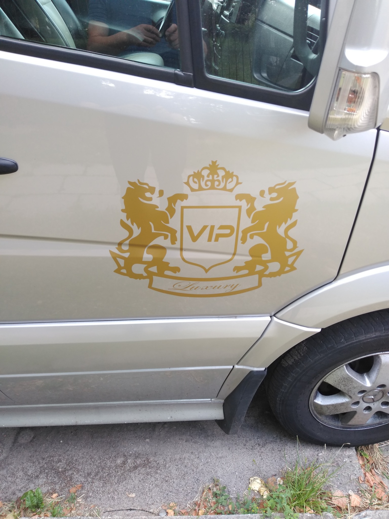 Bok srebrnego vana z naklejką VIP Luxury z dwoma lwami i koroną, widoczny fragment koła i kierowca w odbiciu szyby.