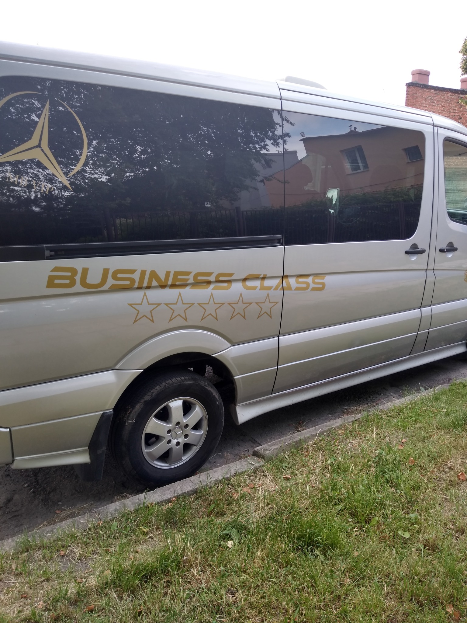 Srebrny bus Mercedes Sprinter z napisem 'BUSINESS CLASS' i pięcioma złotymi gwiazdkami, zaparkowany obok chodnika z widocznym trawnikiem.