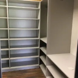 Szara zabudowa szafy wnękowej z półkami i drążkiem na ubrania, widoczna na tle drewnianej podłogi i płyty OSB.