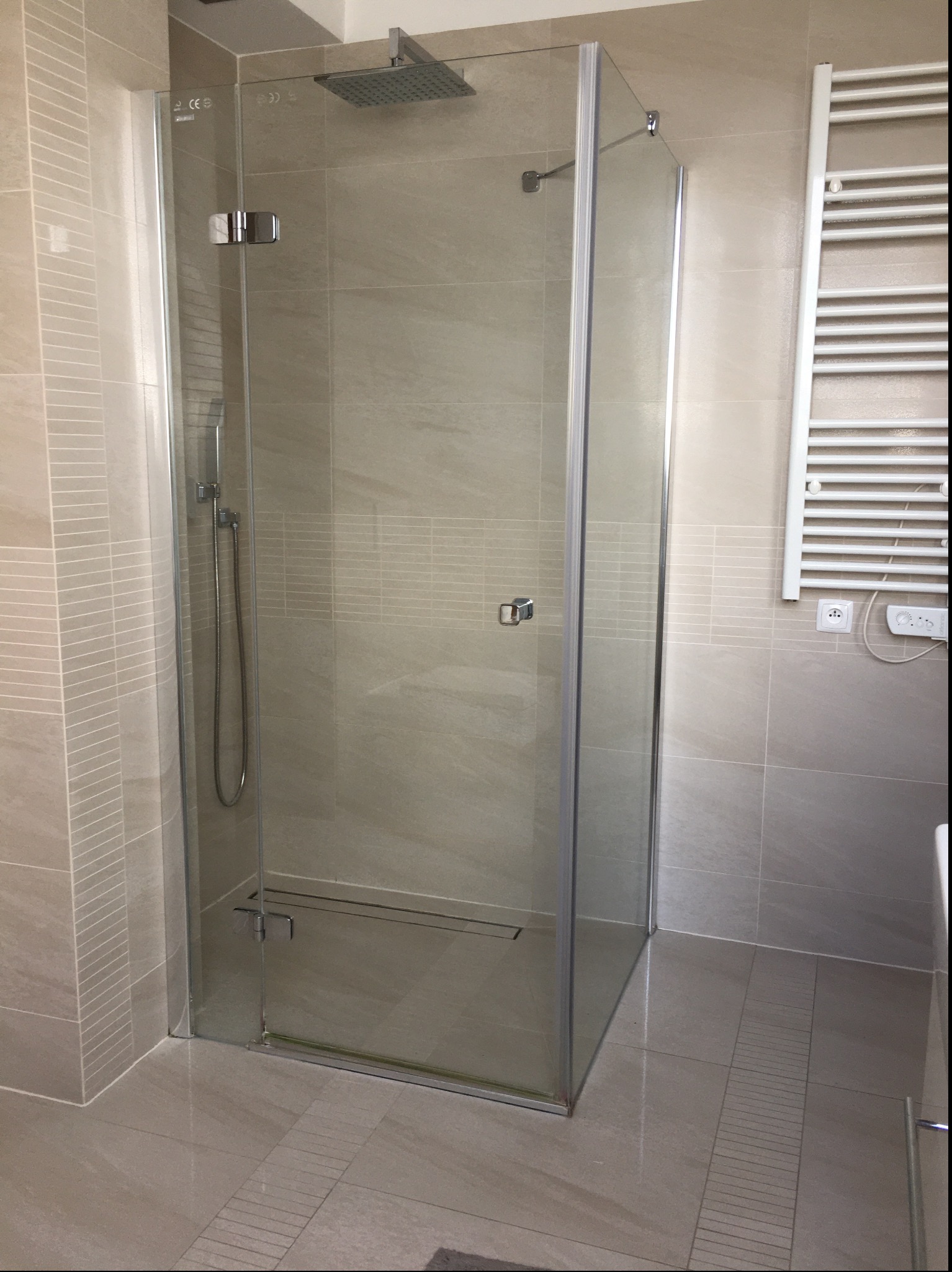 Nowoczesna kabina prysznicowa z transparentnego szkła, chromowane okucia, deszczownica i odpływ liniowy w podłodze wyłożonej beżowymi płytkami, widoczny grzejnik drabinkowy.