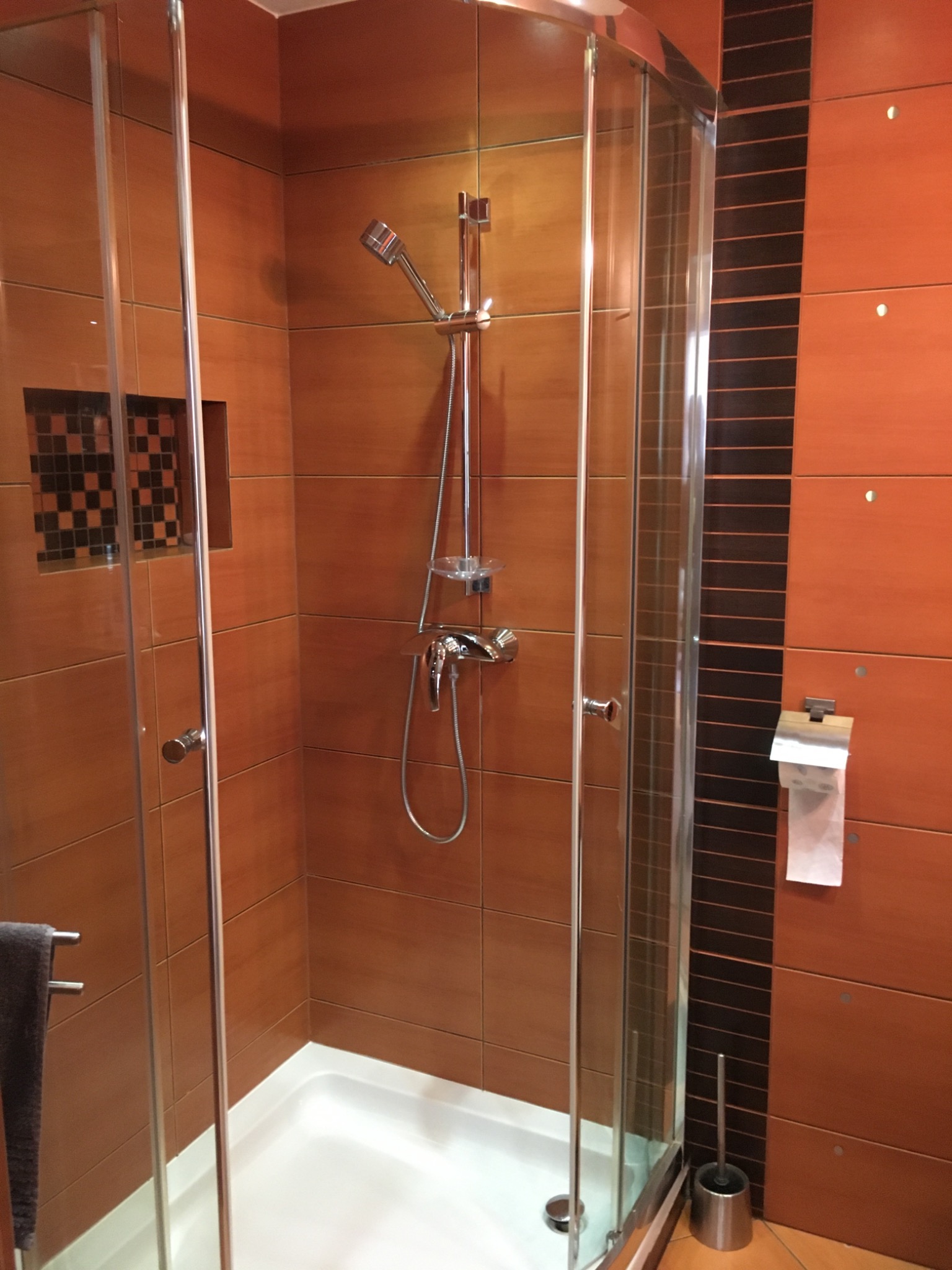 Kabina prysznicowa z brązowymi płytkami imitującymi drewno, czarna mozaika w niszy, chromowana armatura i akcesoria, biały brodzik.
