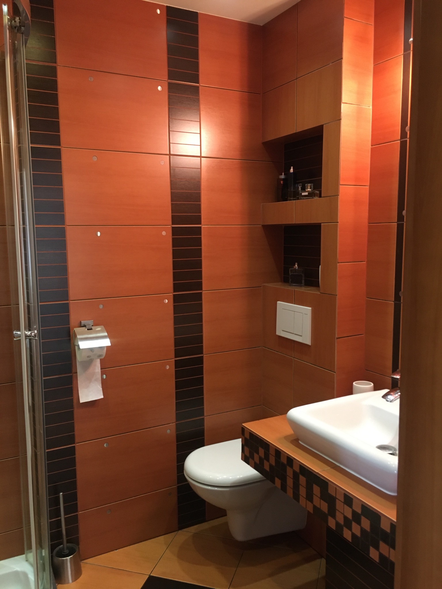 Pomarańczowo-brązowa łazienka z kabiną prysznicową, toaletą, umywalką nablatową i wnękami na kosmetyki, wykończona kwadratowymi płytkami z czarnymi akcentami.