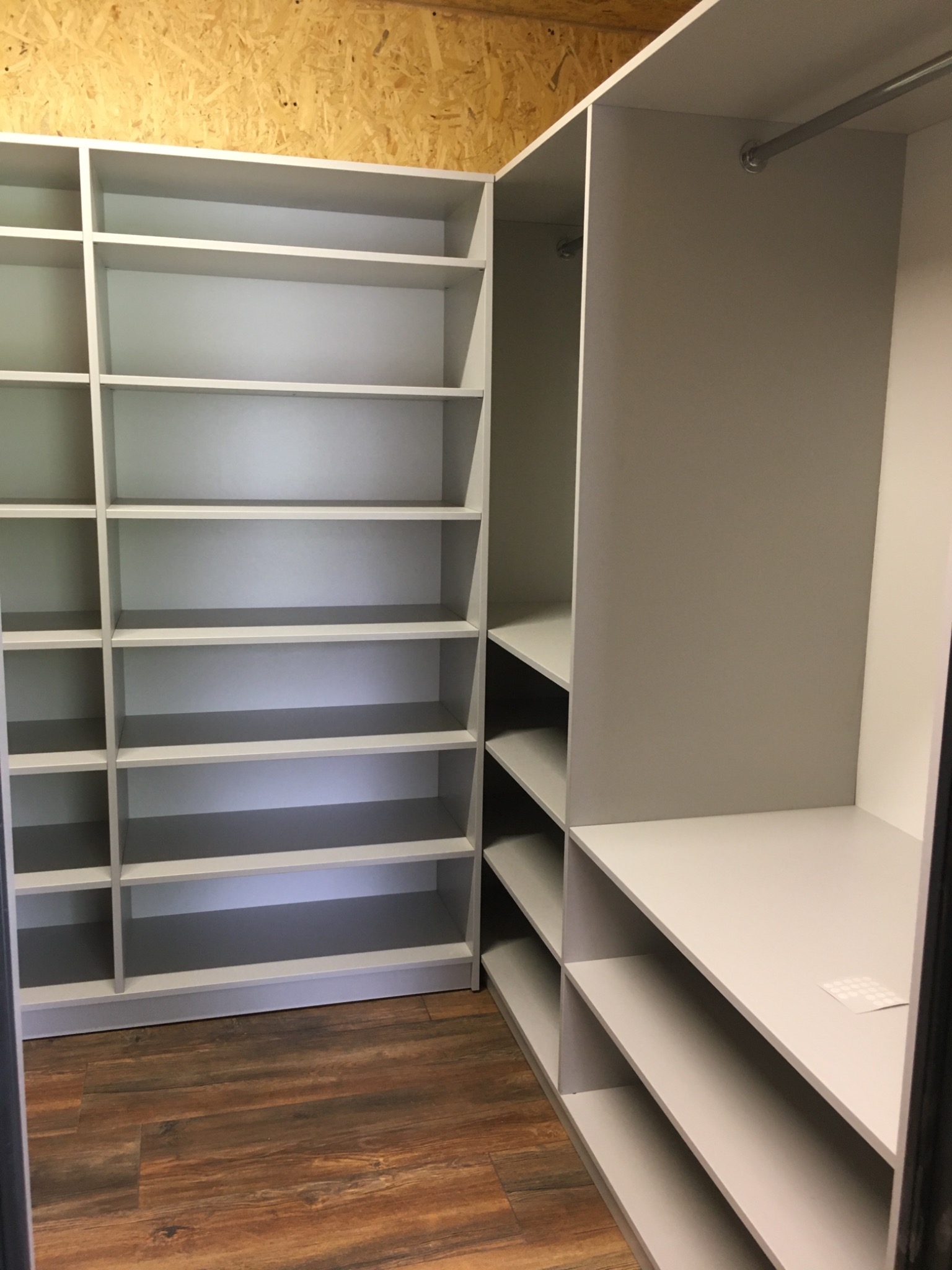 Szara zabudowa szafy wnękowej z półkami i drążkiem na ubrania, widoczna na tle drewnianej podłogi i płyty OSB.