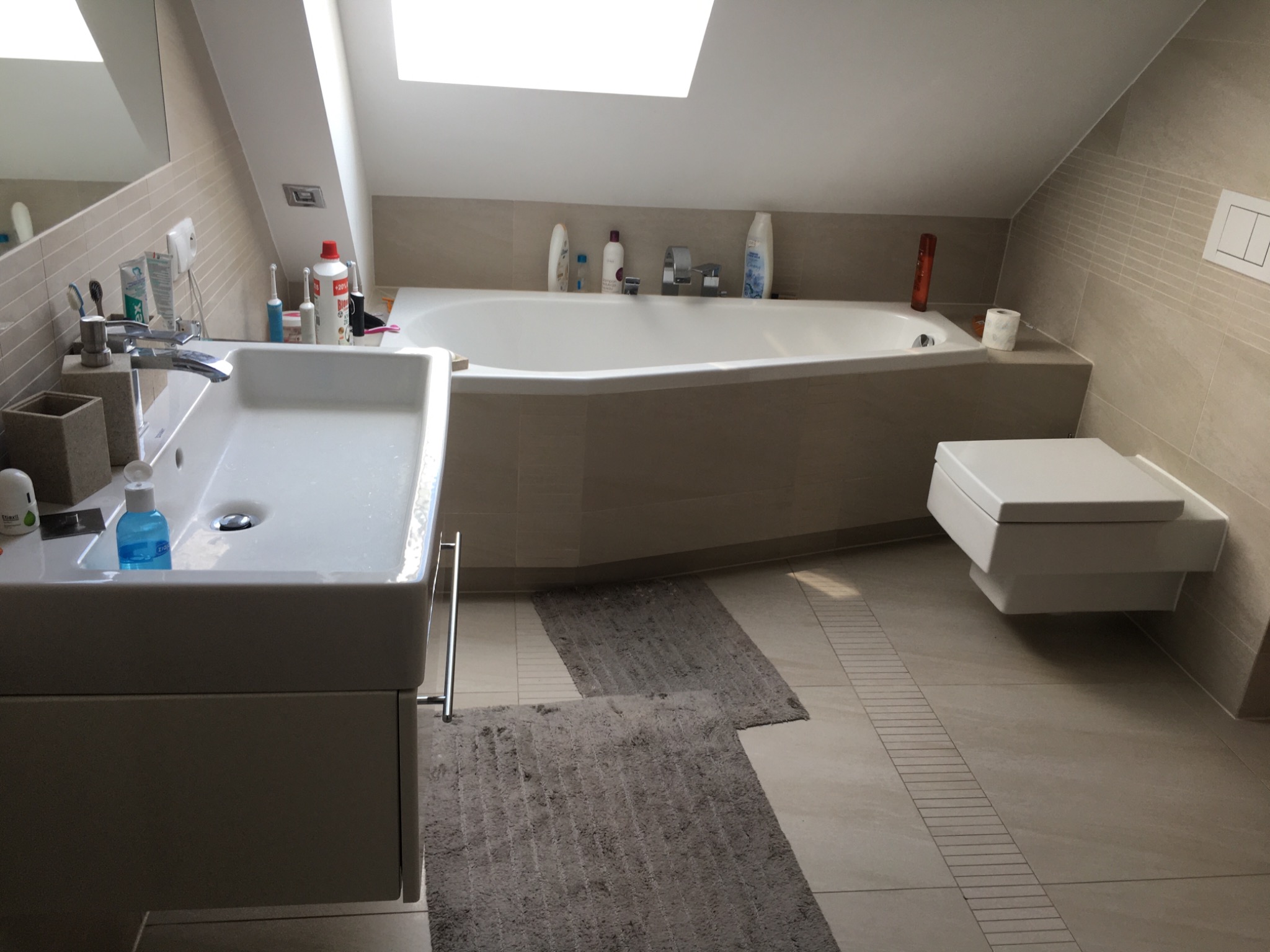 Jasna łazienka na poddaszu ze skośnym sufitem, z wanną obudowaną płytkami w kolorze ścian, wiszącą toaletą i umywalką z szafką, oświetlona światłem dziennym z okna dachowego.