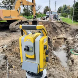Żółto-szary tachimetr Trimble na statywie podczas prac ziemnych, w tle koparka Liebherr i wykop z instalacjami.