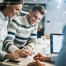 Uśmiechnięta para podpisuje dokumenty przy biurku w jasnym pomieszczeniu biurowym, obok laptop z widocznym tekstem, widok przez szklaną ścianę.