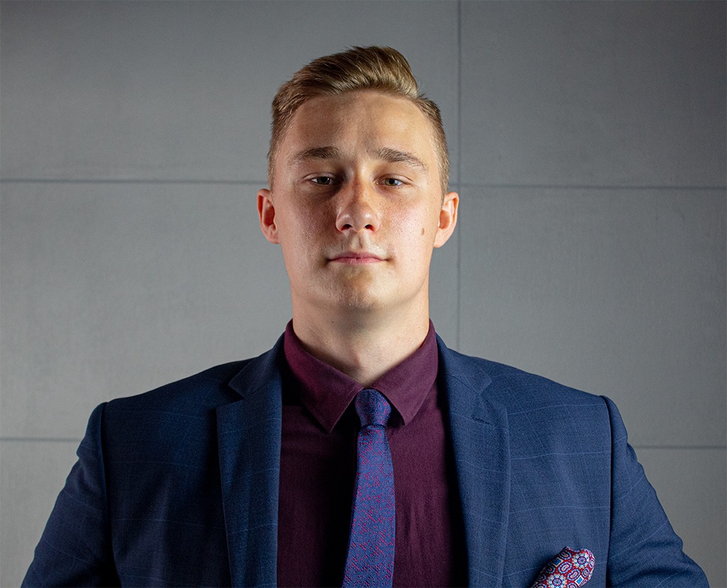 Portret młodego mężczyzny w granatowym garniturze i bordowej koszuli, z wzorzystym krawatem i poszetką, na tle szarej ściany.