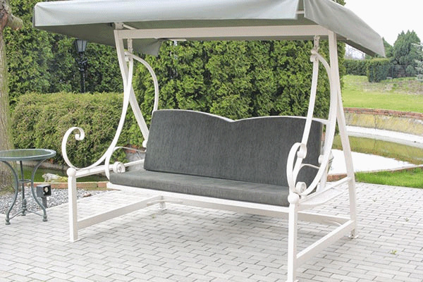 Biała, metalowa huśtawka ogrodowa z szarymi poduszkami i daszkiem, stojąca na brukowanym patio, z zielonymi krzewami i małym stawem w tle.