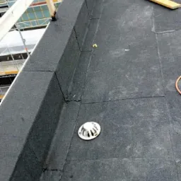 Wykończona, czarna, płaska powierzchnia dachu z widocznym odpływem liniowym i fragmentem konstrukcji rusztowania w tle.