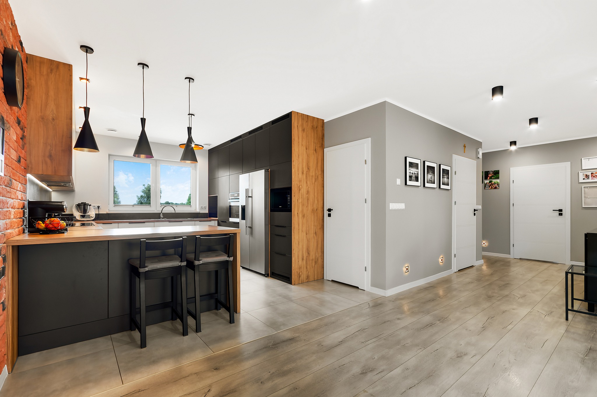 Przestronna kuchnia z wyspą, czarnymi szafkami i drewnianymi akcentami; widoczne wiszące lampy, ceglana ściana i drewniana podłoga.