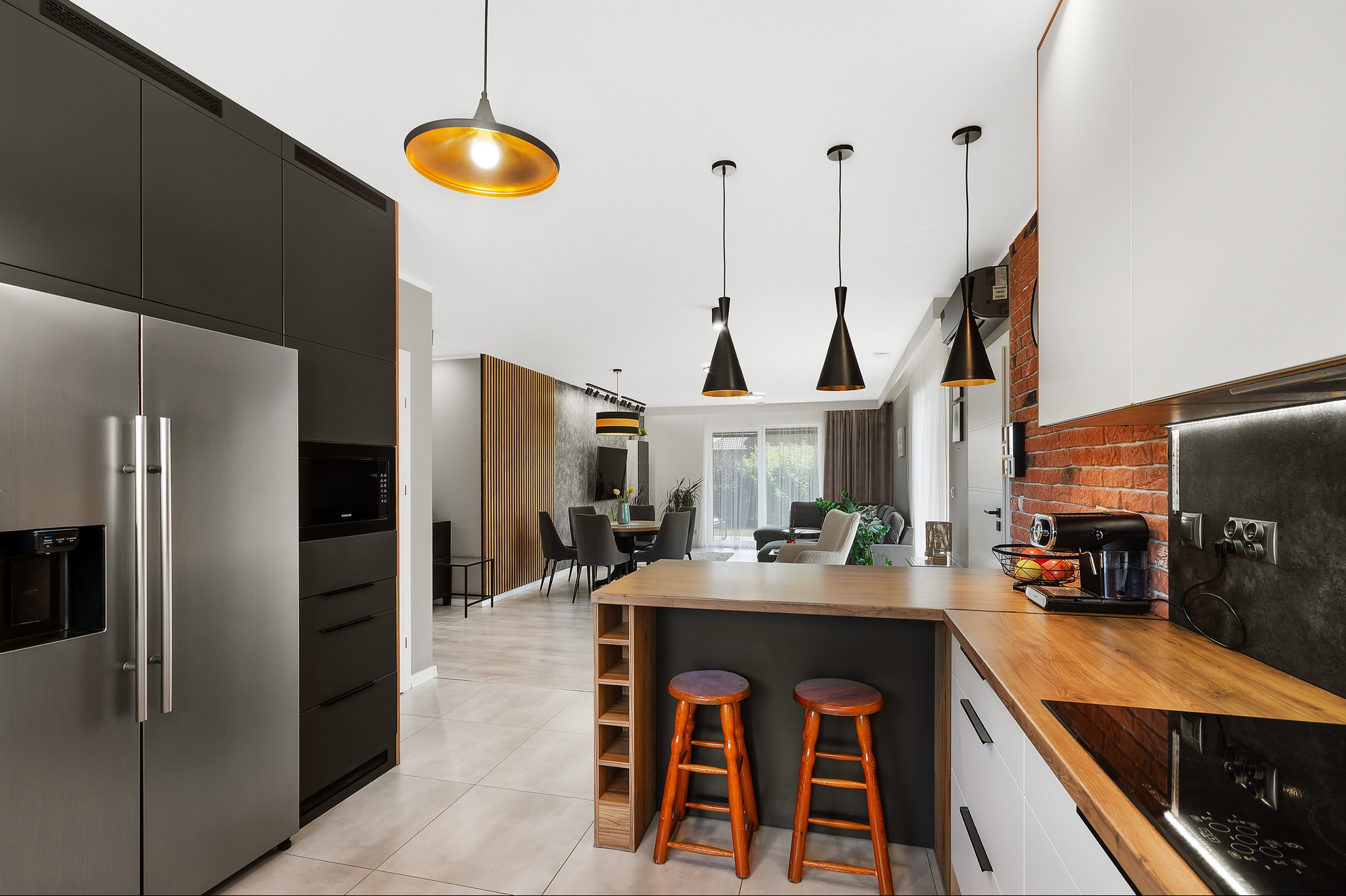 Nowoczesna kuchnia z wyspą i hokerami, widok na salon z jadalnią, czarne lampy wiszące nad blatem, ściana z czerwonej cegły w tle, sprzęt AGD ze stali nierdzewnej.