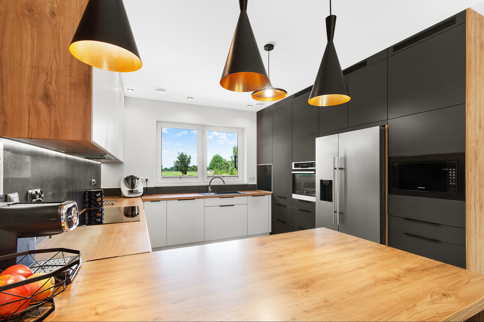 Nowoczesna kuchnia z drewnianym blatem wyspowym, czarnymi wiszącymi lampami z miedzianym wnętrzem, białymi szafkami i zabudową w kolorze grafitowego drewna z wbudowanym sprzętem AGD.
