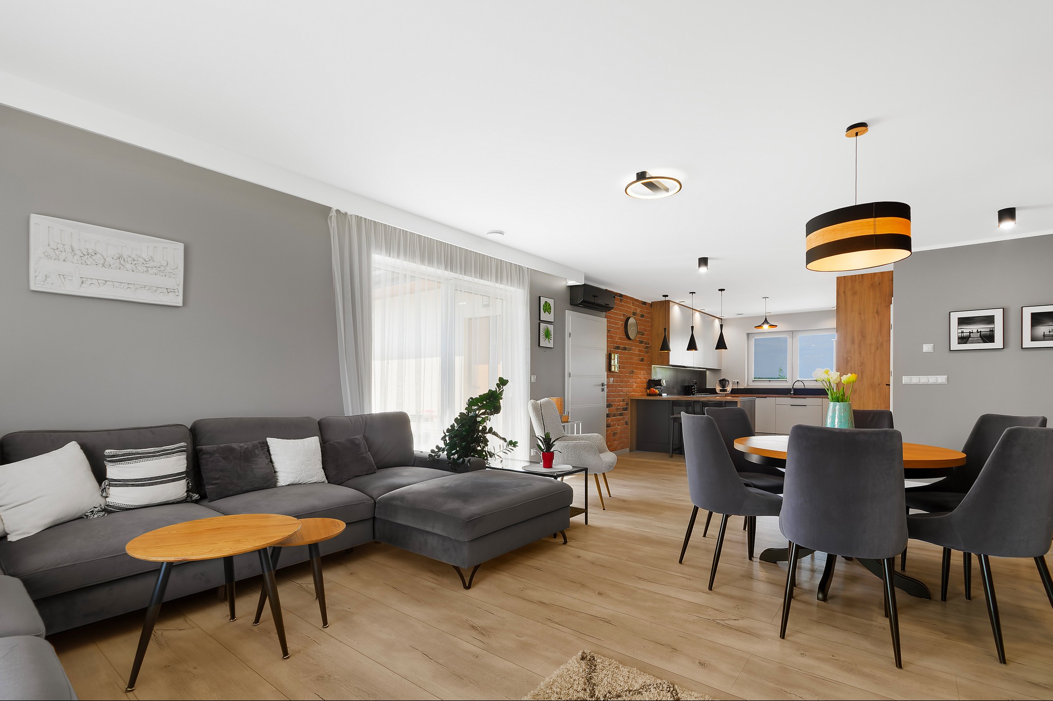 Przestronny salon z aneksem kuchennym w Biała Podlaska, szara sofa narożna z drewnianymi stolikami kawowymi, okrągły stół z szarymi krzesłami, ceglana ściana w tle.