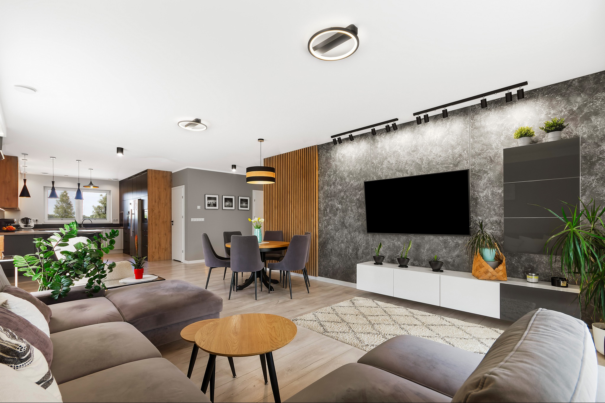 Przestronny salon z szarą sofą, okrągłym drewnianym stolikiem kawowym, stołem z krzesłami, ścianą TV z szarą strukturą i oświetleniem punktowym oraz widokiem na kuchnię.