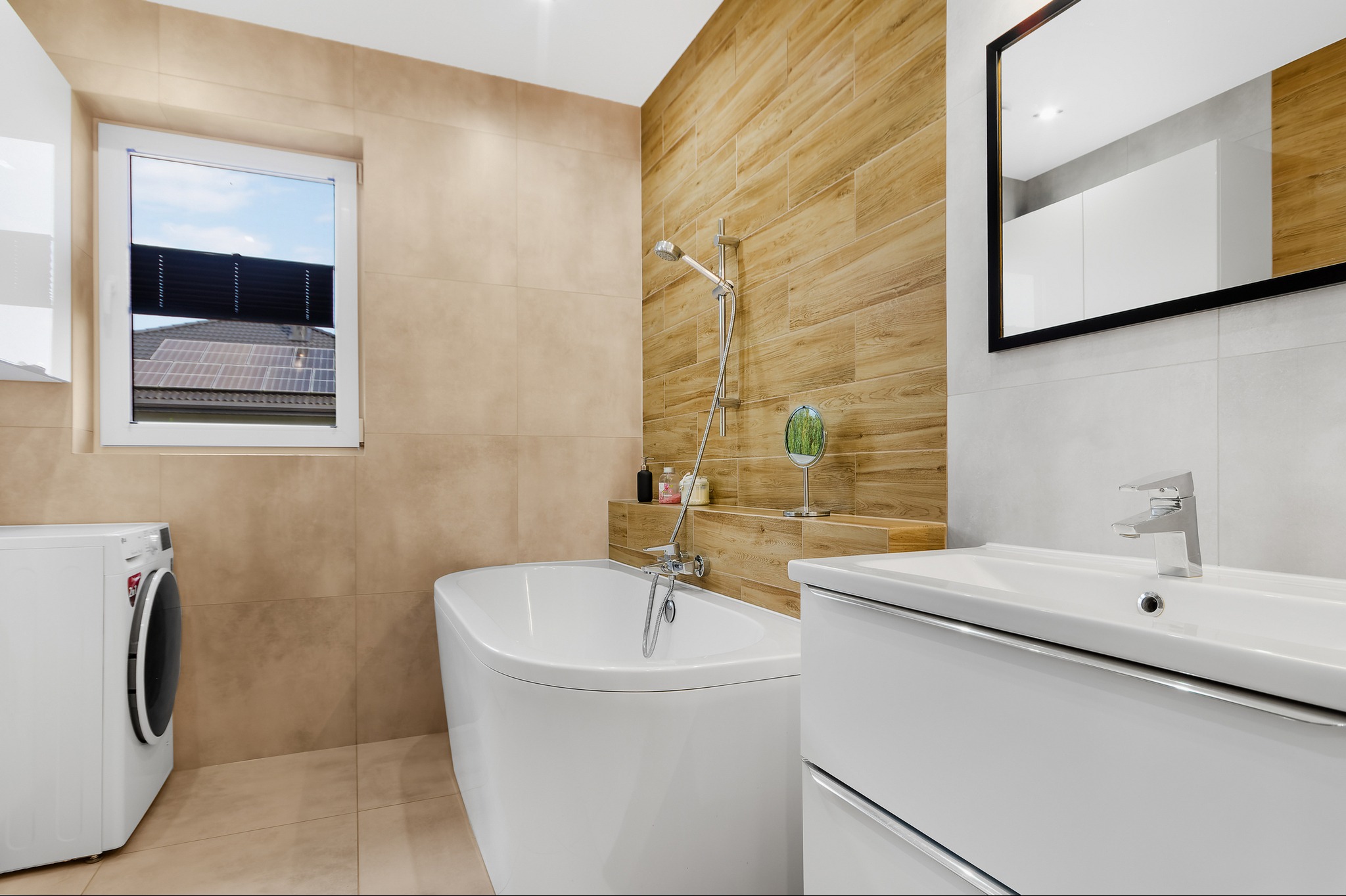 Nowoczesna łazienka z wanną wolnostojącą, pralką, umywalką z szafką i oknem z widokiem na dach z panelami słonecznymi.