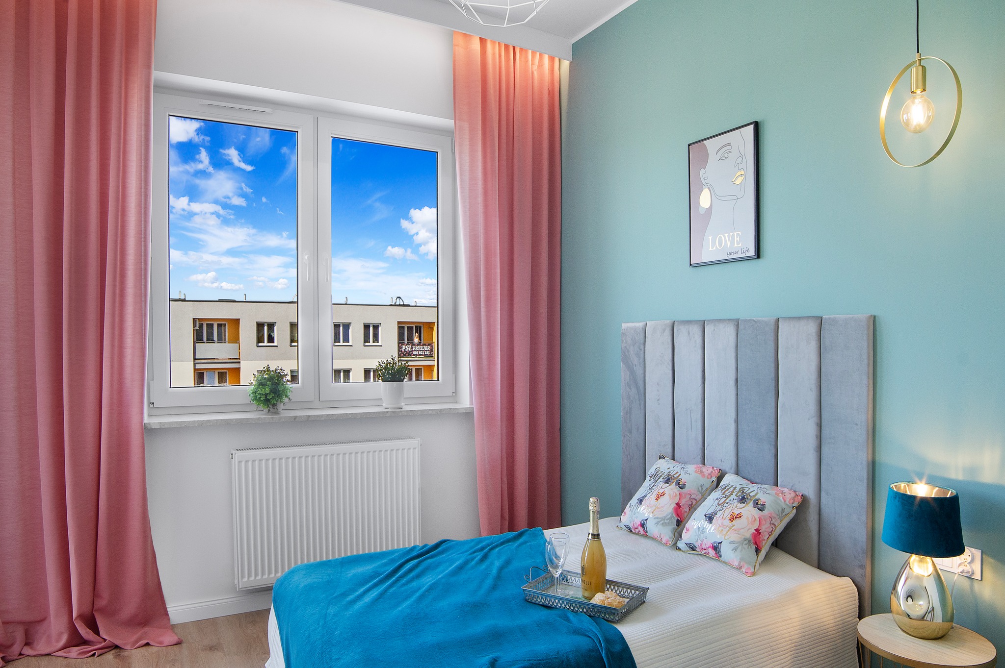 Jasne wnętrze sypialni z turkusowymi ścianami, szarą tapicerowaną ramą łóżka, niebieską narzutą i tacą z szampanem, widokiem na błękitne niebo przez okno z różowymi zasłonami.