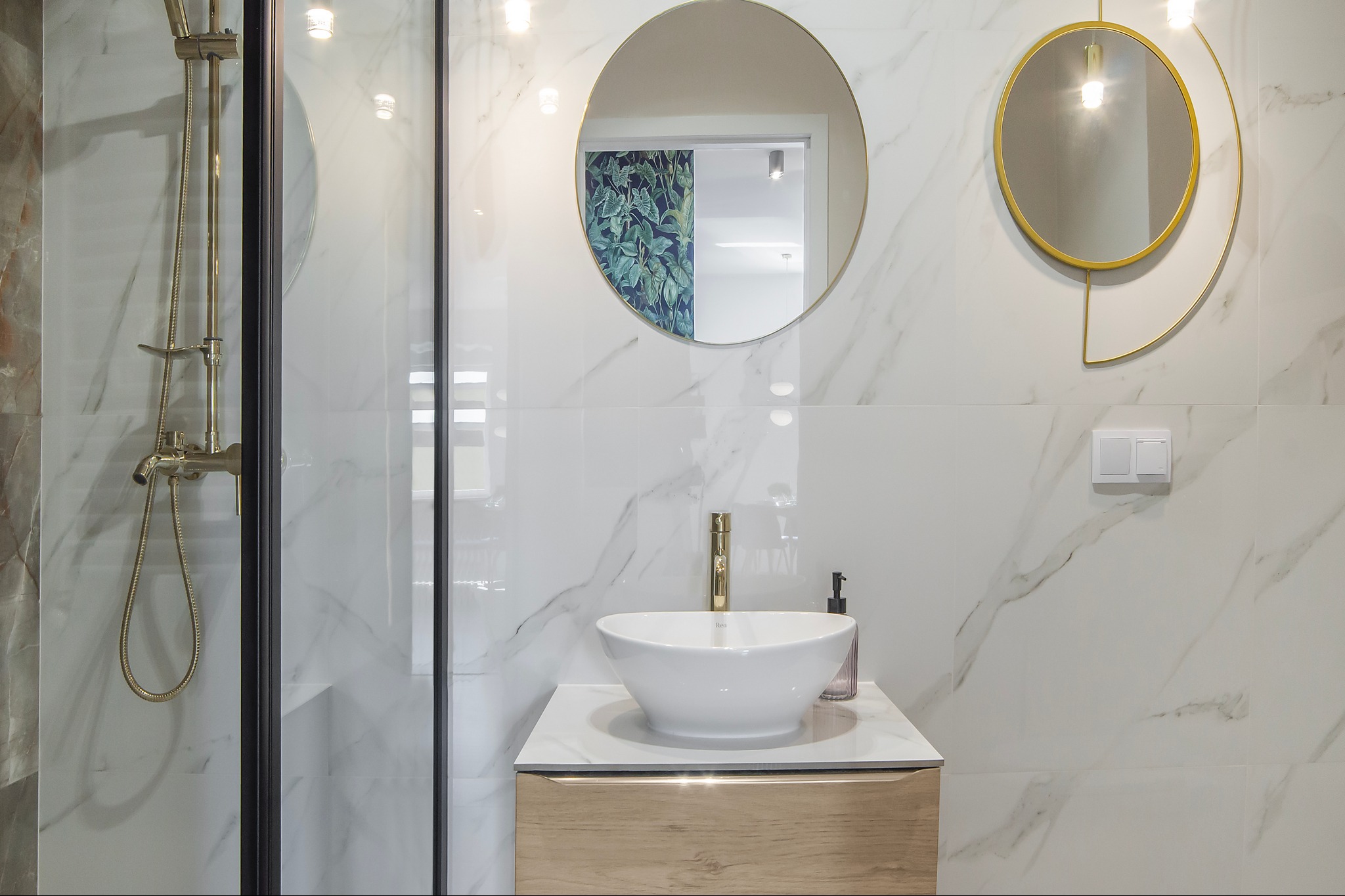Nowoczesna łazienka z białymi marmurowymi płytkami, okrągłymi lustrami w złotych ramach, umywalką nablatową i złotą armaturą pod prysznicem z czarną ramą kabiny.