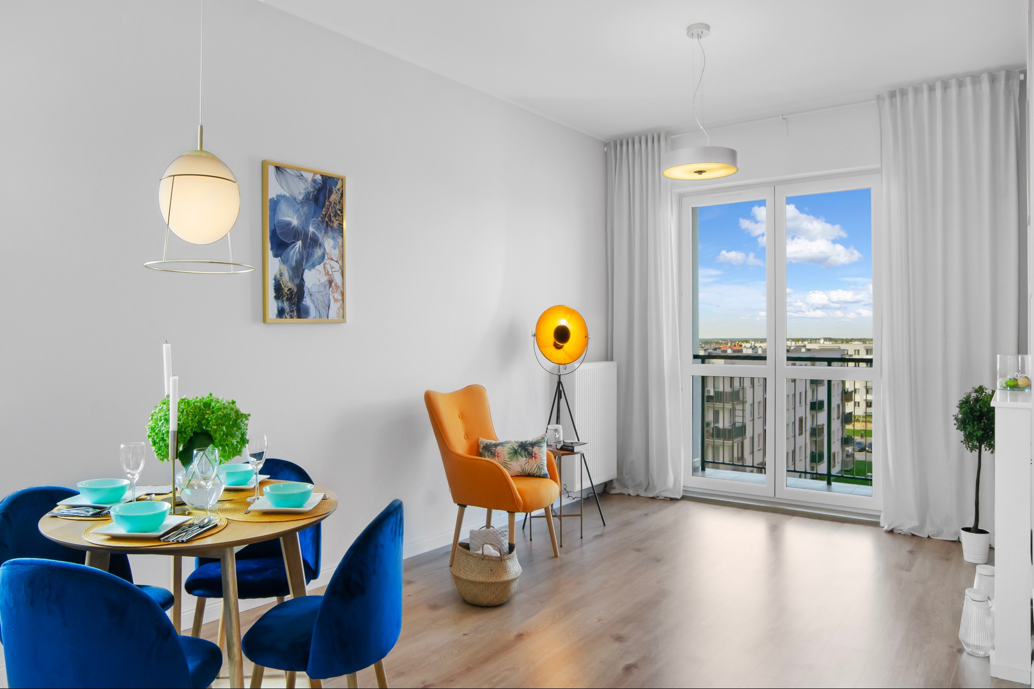 Jasne wnętrze mieszkania z okrągłym stołem jadalnym, aksamitnymi krzesłami, pomarańczowym fotelem i lampą studyjną na trójnogu, widok na miasto z balkonu.