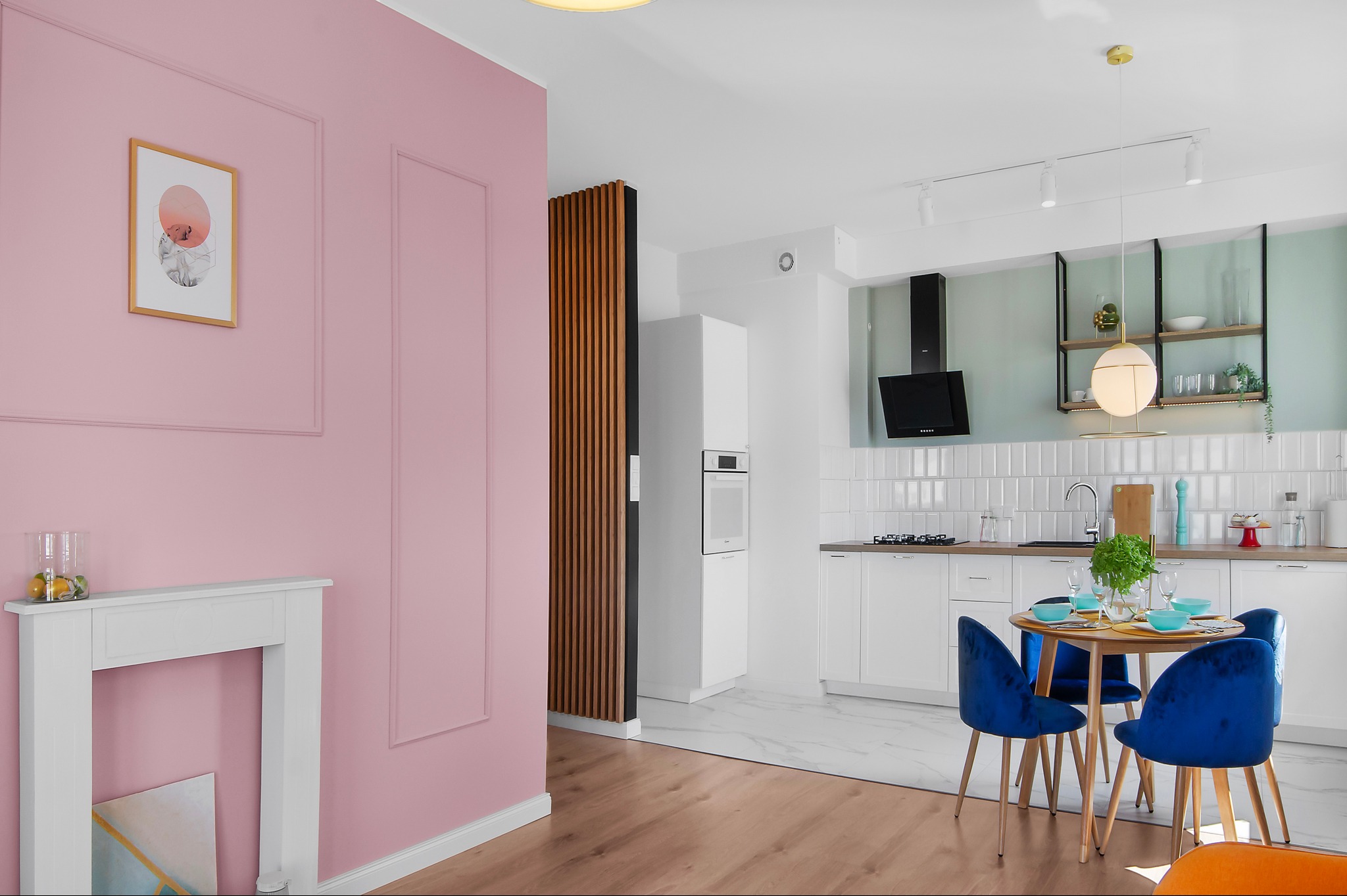Jasne wnętrze: różowa ściana z dekoracyjną ramą, biały kominek, okrągły stół z czterema niebieskimi krzesłami, biała kuchnia z czarnym okapem i marmurową podłogą, całość oświetlona nowoczesnymi...