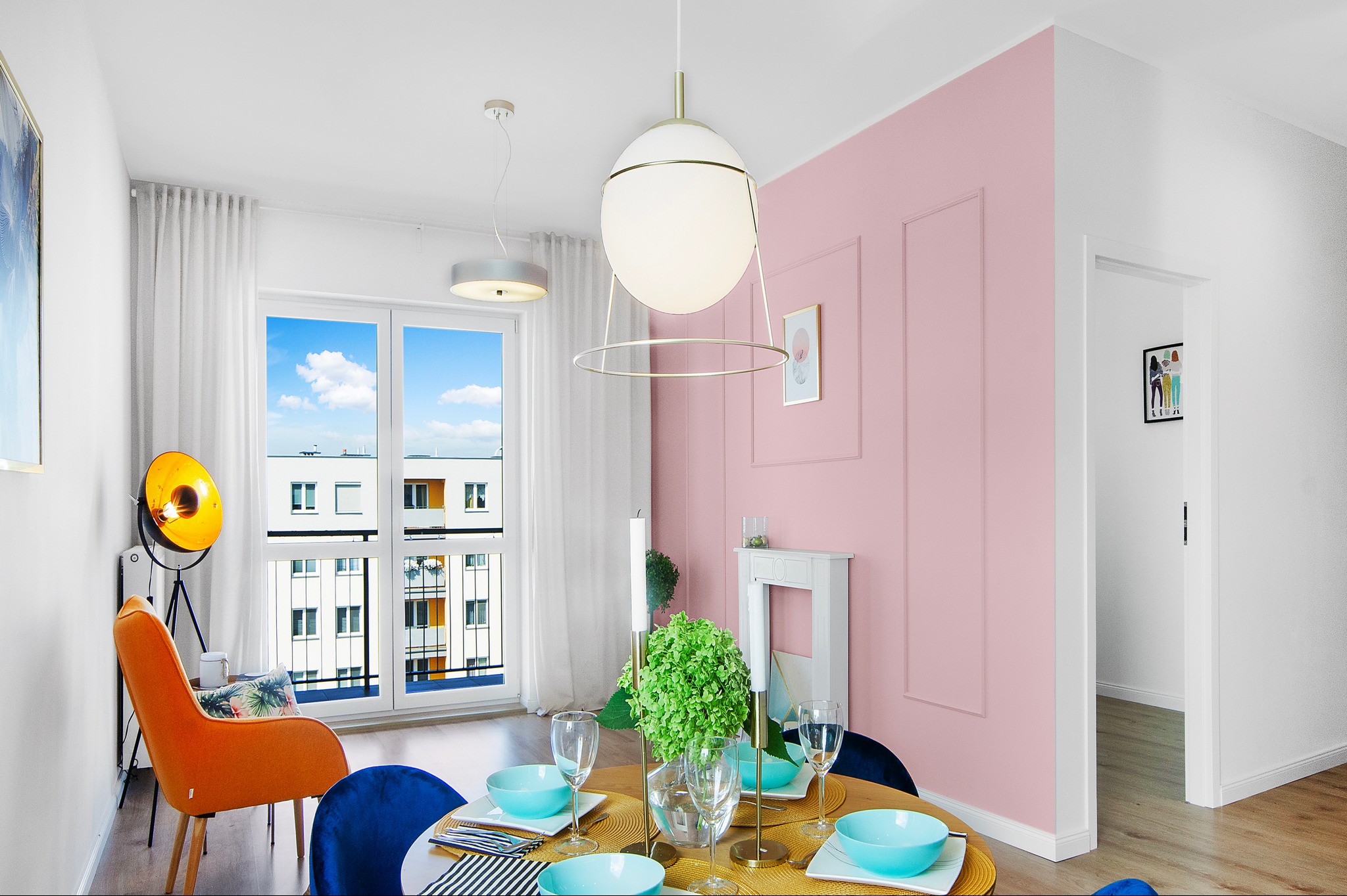 Jadalnia w pastelowych kolorach z okrągłym stołem, niebieskimi miseczkami i złotymi podkładkami, różowa ściana z dekoracyjnymi listwami, widok na budynki przez okno.