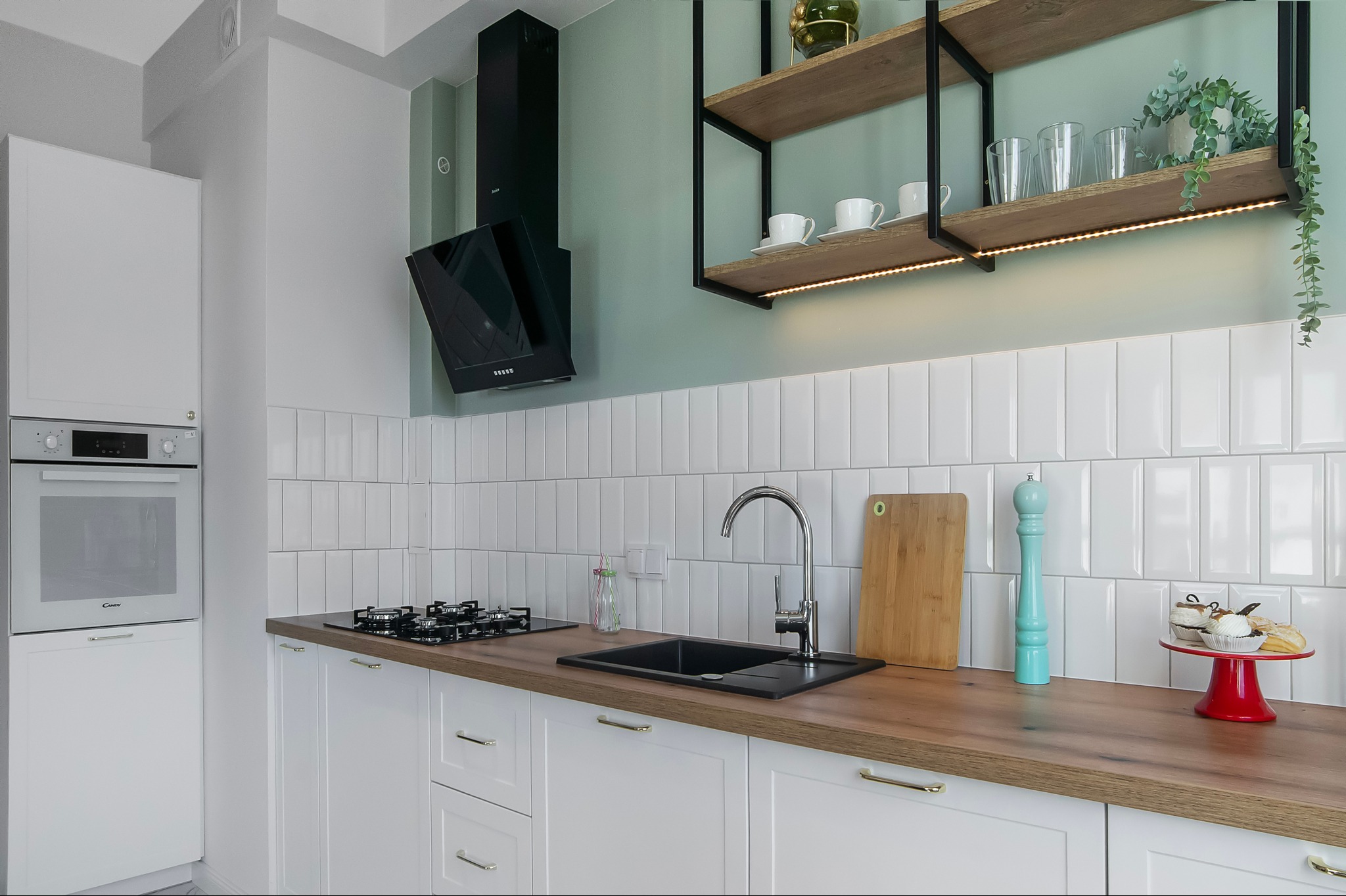 Nowoczesna kuchnia z białymi szafkami, drewnianym blatem, czarnym zlewem i płytą gazową, uzupełniona o drewniane półki z oświetleniem LED i dekoracjami.