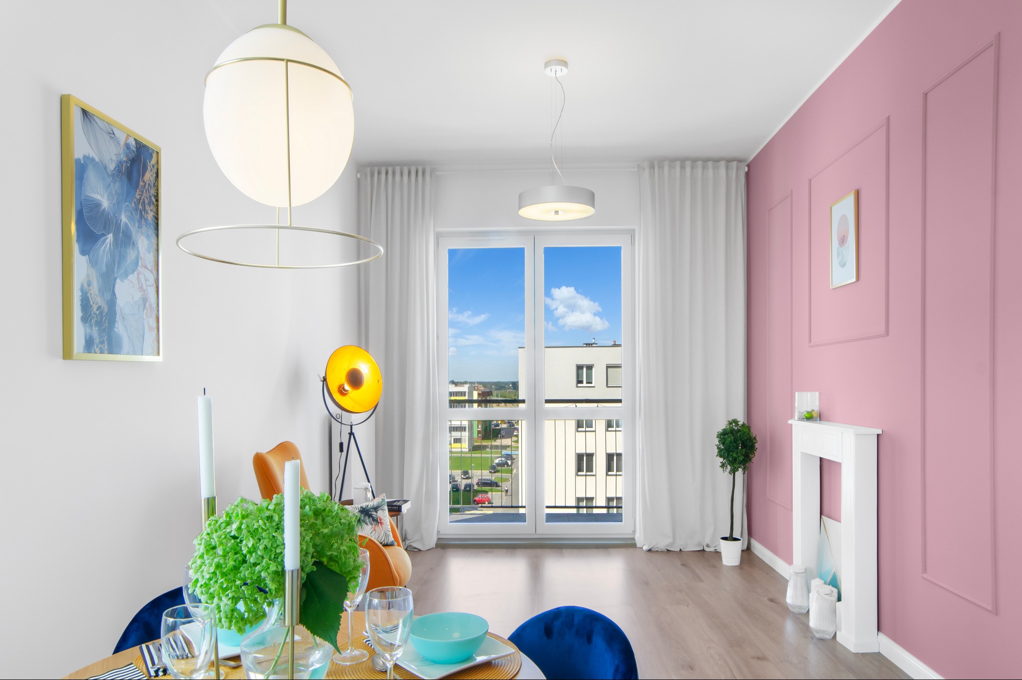 Jasne wnętrze z różową ścianą, ozdobnym kominkiem, okrągłym stołem z niebieskimi krzesłami i widokiem na budynek mieszkalny za oknem.