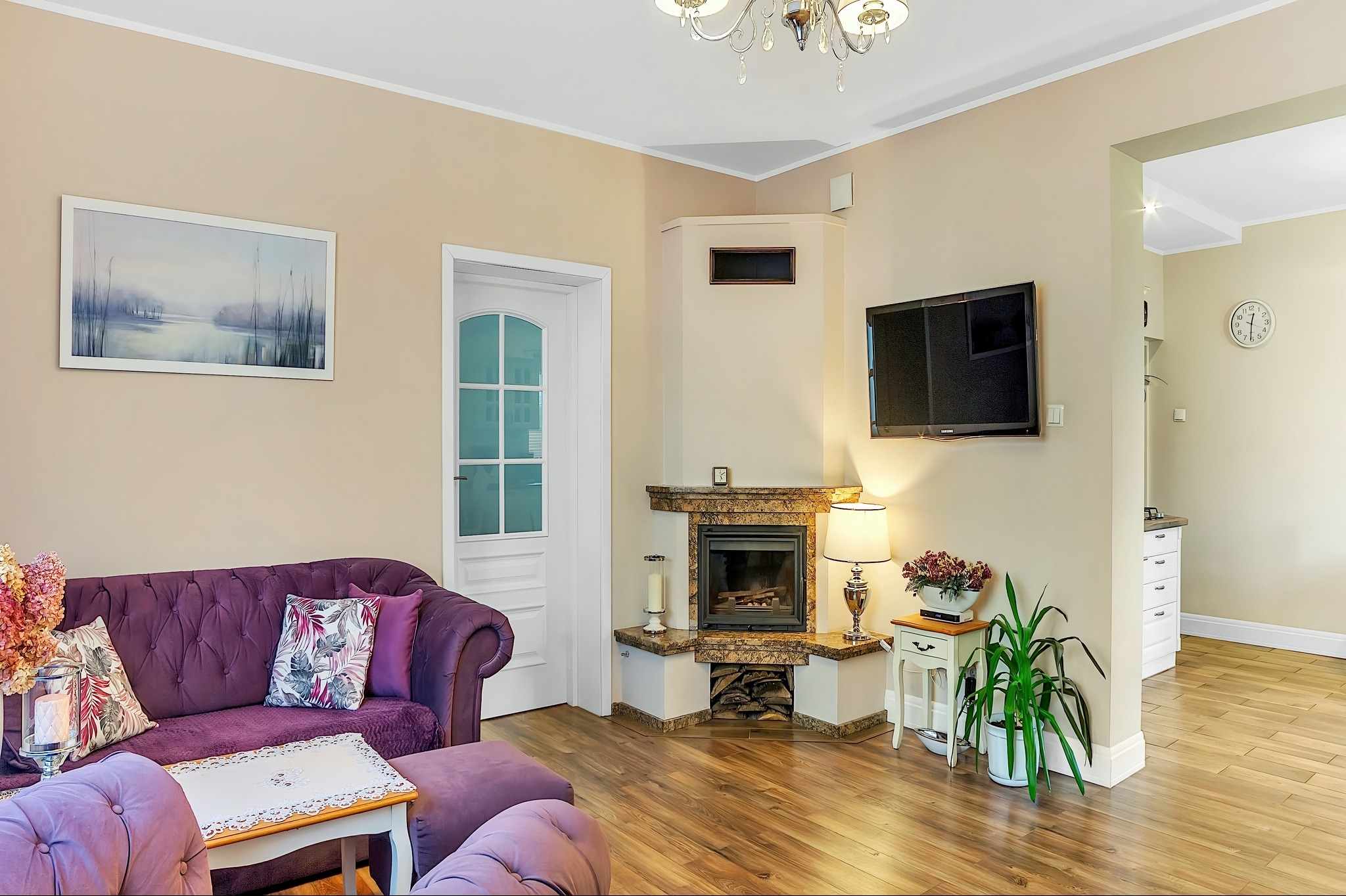 Salon z fioletową sofą Chesterfield, marmurowym kominkiem z paleniskiem, telewizorem na ścianie i widokiem na kuchnię z białymi szafkami.